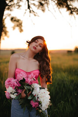 Naklejka premium Stylish girl with long wavy hair holds a bouquet of peonies in the field at sunset.