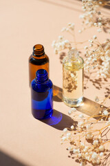 Open amber, blue and transparent bottles without cap with serum or essential oil with white flowers. Beige background with daylight and beautiful shadows. Beauty concept for face and body care