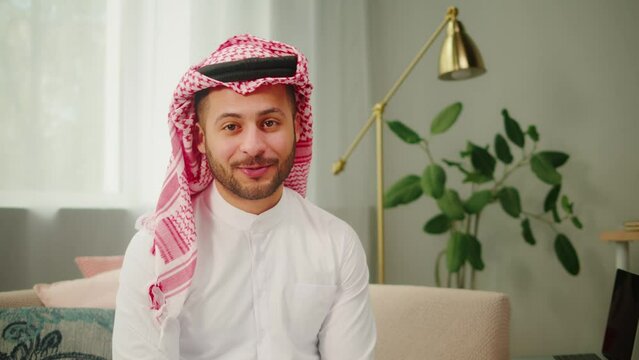 Middle eastern man using virtual conference. Male person in earphones talking on video call in living room. Wearing traditional Islamic clothes. Communicating with family and friends. 