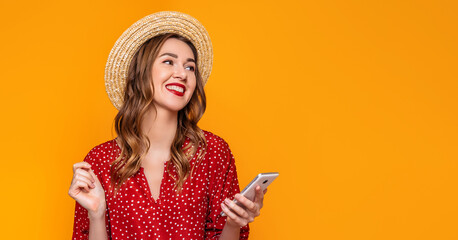 Young stylish girl in a red vintage dress, a straw hat, with red lipstick smiles, laughs, listens to music on the mobile phone and looks at copy space isolated on a yellow, an orange background