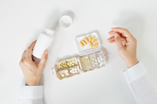 Pills box for daily intake of vitamins and medicines on white table. Top view different capsules and tablets in open plastic container in woman hands. Lifestyle for beauty and health, flat lay
