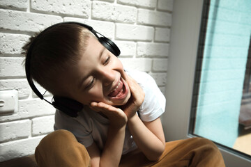 A boy of 6-7 years old in a white T-shirt listens to music in black headphones and sings, smiles