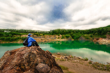 Asian traveller man with backpack sitting on the rock in nature pond water
