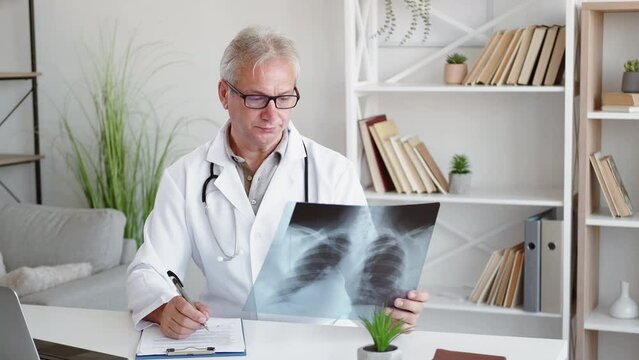 Lung diagnostic. Medical examination. Obstructive disease. Pensive puzzled middle-aged male doctor in glasses studying chest xray scan film at modern workplace interior.