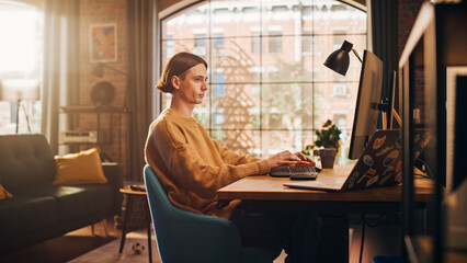 Fototapeta na wymiar Young Handsome Man Working from Home on Desktop Computer in Sunny Stylish Loft Apartment. Creative Designer Wearing Cozy Yellow Sweater and Headphones. Urban City View from Big Window. Static Shot.