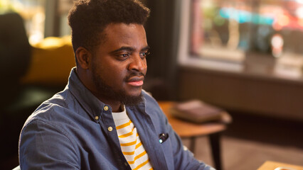 Fototapeta premium Young Handsome Black Man Working from Home on Desktop Computer in Sunny Stylish Loft Apartment. Creative Male Checking Social Media, Browsing Internet. Urban City View from Big Window.