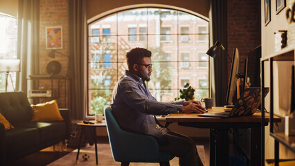 Fototapeta na wymiar Young Handsome Black Man Working from Home on Desktop Computer in Sunny Stylish Loft Apartment. Creative Male Checking Social Media, Browsing Internet. Urban City View from Big Window.