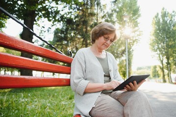 Attractive mature woman sitting on a bench holding and using a digital tablet in the park at summer day