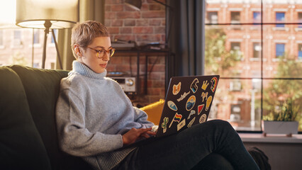 Young Attractive Woman Working from Home on Laptop Computer with Stickers in Sunny Stylish Loft Apartment. Creative Designer Wearing Cozy Blue Sweater and Glasses. Urban City View from Big Window. - obrazy, fototapety, plakaty