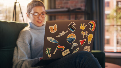 Stylish Female Using Laptop Computer with Diverse LGBT and Lifestyle Stickers on the Back. Young Creative Woman Sitting on a Couch, Typing, Browsing Internet and Checking Social Media at Home. - obrazy, fototapety, plakaty
