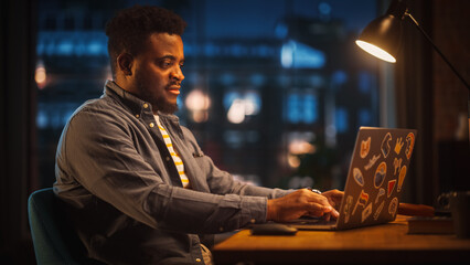 Young Handsome Black Man Working from Home on Laptop Computer in Stylish Loft Apartment in the Evening. Creative Male Gets a Great Idea while Checking Social Media. Urban City View from Big Window.