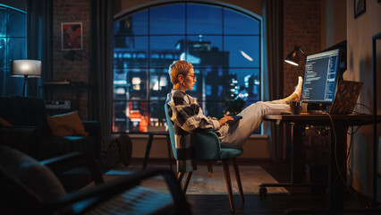 Young Woman Writing Code on Computer in Stylish Loft Apartment in the Evening. Creative Female Wearing Cozy Clothes, Resting Feet on Table and Using Wireless Keyboard. Urban City View from Big Window.