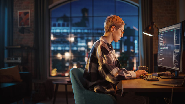 Young Woman Writing Code On Desktop Computer In Stylish Loft Apartment In The Evening. Creative Female Wearing Cozy Clothes, Working From Home On Software Development. Urban City View From Big Window.