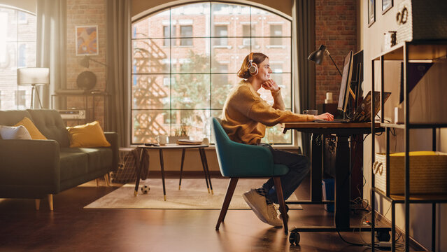 Young Handsome Man Working from Home on Desktop Computer in Sunny Stylish Loft Apartment. Creative Designer Wearing Cozy Yellow Sweater and Headphones. Urban City View from Big Window.