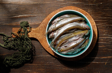 Morning catch. Small raw fish on a vintage wooden board and a wicker string bag. Top view.