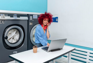 a smiling young latin woman with a red afro hair works with her laptop and talks on the phone in the blue laundry room, while waiting for the laundry to be done, her red headphones around her neck - Powered by Adobe