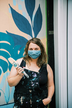 Woman With Mask Standing In Front Of Mural With A Paint Brush