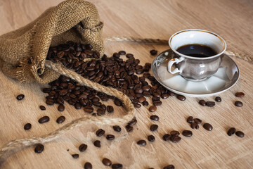 Roasted coffee beans in little bag with ropes and cup of coffee with drink on dark brown wooden surface, in the morning for breakfast.