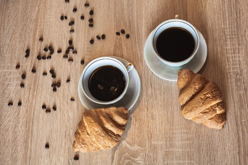 Two cup of hot coffee in a plate and a croissants. Coffee beans are scattered on a wooden light brown table.