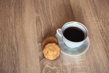 A cup of hot fragrant black coffee in a saucer with cookies, on a wooden light brown textured table.