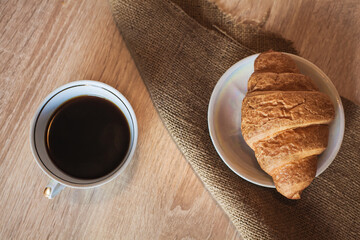 A cup of hot fragrant black coffee with a croissant in a saucer on a cloth, on a light brown wooden table.
