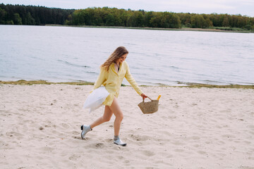 A girl with blond curly hair, dressed in a yellow shirt, with a basket and a pillow in her hands, walks and smiles, on the beach by the river. The basket contains a bottle of champagne.