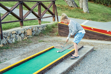 Boy enthusiastically plays mini golf on summer evening on the territory of the hotel
