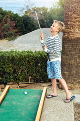 Boy enthusiastically plays mini golf on summer evening on the territory of the hotel - 511097334