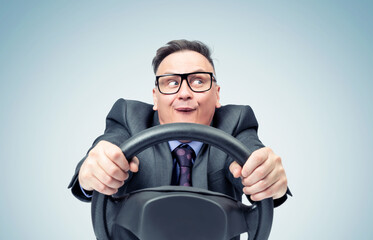 Portrait of a comical businessman in a dark suit and glasses holding a car steering wheel in his...