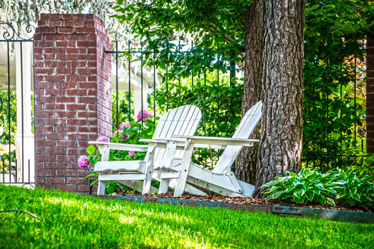 Two white wooden Adirondack chairs sit by elegant iron fence with Hydrangea flowers in backgound under trees