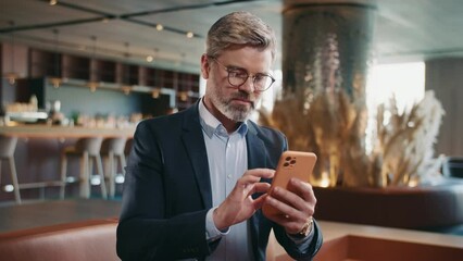 Shot of handsome smiling man in formal outfit using smartphone in restaurant, touching screen of mobile phone, scrolling social media. Communication. Indoors. Business people. Working day. Summertime - Powered by Adobe