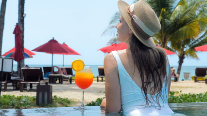 Fototapeta premium Woman in white swimwear and hat relax in swimming pool enjoy beautiful sea view with sun beds at sandy beach with tropical palms. Female traveler sunbathing in resort with glass of orange mocktail