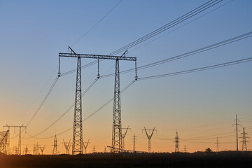 Dark silhouettes of power lines on fiery sunrise. High voltage electricity towers in field and fiery sunset. Concept of crisis of energy consumption, generation, and supply.