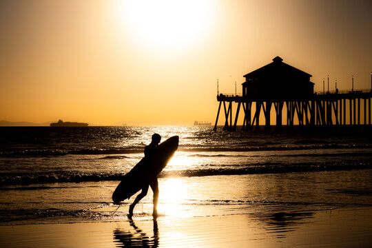 Surfer Walking on Beach Carrying Surfboard in Front of the Pier at Huntington Beach California at Sunset
