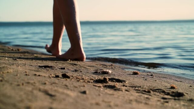 Slow motion close-up view of a barefoot feet of a young woman walk on the sand along the coast at sunset. Summer vacation holiday.
