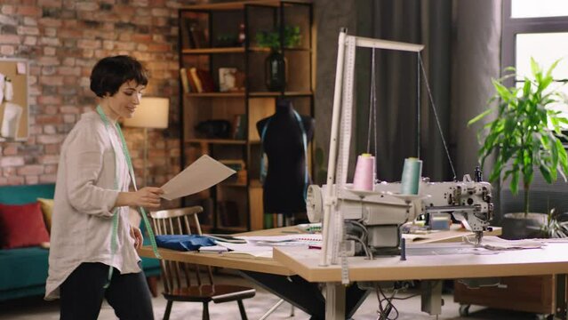 Concept of small business tailoring fashion people talented dressmaker woman with a pretty smile take her sketches from the work table came to the sew mach.ine and start to sewing some fabrics. Shot