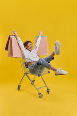 Happy girl holding colorful packages, while riding in shopping cart indoor. Side view of female buyer having fun in shop trolley, while shopping, isolated on orange background. Concept of happiness.
