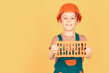 Boy in a construction helmet holds a brick in his hands on yellow background. Little builder in...