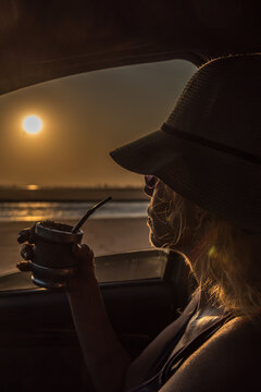 Woman inside a car with a hat drinking mate looking at the sunset over the sea
