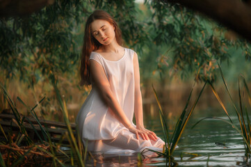 A slender brunette with long hair in a wet transparent dress sits in the water. Gentle portrait of...