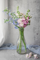 Bouquet of Blue Bellflowers, Pink Stock, and White Hypericum Berries in an Old Milk Bottle near a Window; Concrete Table and Background; Blue Fabric