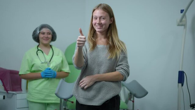 Charming teenage girl looking back at gynecologist and looking at camera gesturing thumb up smiling. Portrait of confident positive Caucasian teen posing in hospital on appointment advertising