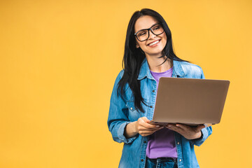 Portrait of happy young beautiful surprised woman with glasses standing with laptop isolated on yellow background. Space for text. - 511121301