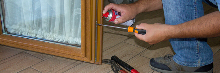 Image of the hands of a handyman as he repairs and spray lubricates the lock of a house window....