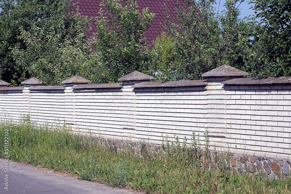 Sticker long white wall of brick fence on brown stone foundation on a rural street in green grass