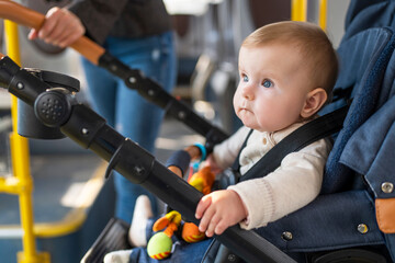 A cute little baby of 10 months is sitting in a stroller. A child is fastened with seat belts in stroller rides a passenger bus with her mother. Concept of child safety.