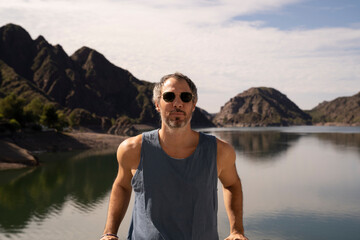 Portrait of a fit man in his 30s, wearing a muscle vest and sunglasses. View of the mountains and sky reflection in the lake water, in the background.
