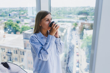 Fototapeta premium Happy woman drinks coffee and looks out the window in a cafe. A young smiling girl in glasses is standing by the window. Recreation, leisure and free time. The modern freelancer lifestyle