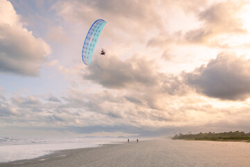 homem voando com seu parapente sobre a praia extensa em final de tarde - obrazy, fototapety, plakaty