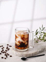 Iced coffee in a glass with cream, ice cubes and grains on a light marble background with morning shadows. The concept of a cold summer drink.  Copy space